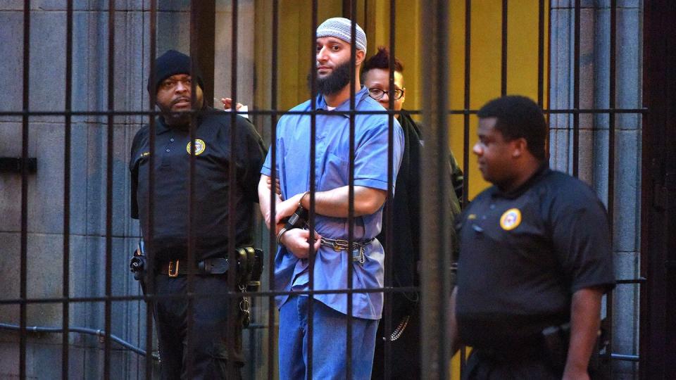 adnan syed wears a blue prison outfit, a gray cap, and handcuffs, he is looking toward the camera from behind bars and three officers are in the vicinity