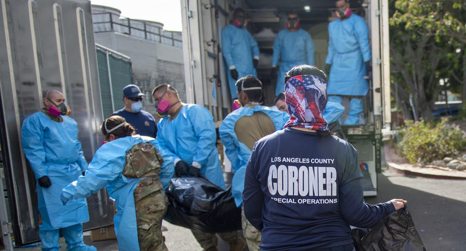 Coroner stands in front of a truck while people in PPE load a body onto to it.