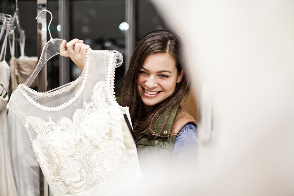 A woman smiles while holding up a white lace dress on a hanger in what appears to be a clothing store