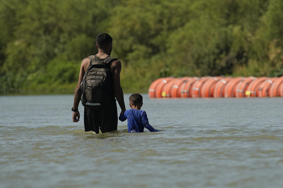 FILE - Migrants walk past large buoys being used as a floating border barrier on the Rio Grande Tuesday, Aug. 1, 2023, in Eagle Pass, Texas. The 5th U.S. Circuit Court of Appeals heard arguments on the future of the barrier of giant buoys that aimed at deterring migrant traffic on Wednesday, May 15, 2024. (AP Photo/Eric Gay, File)