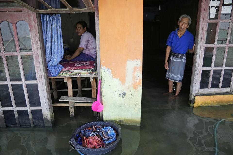 Sudarto stands at the door of his flooded home as his daughter Turiah looks on at their flooded house in Timbulsloko, Central Java, Indonesia, Sunday, July 31, 2022. With a physical disability that prevents Turiah from normal work in the village, she spends her day sitting in the home's front window on an elevated wooden platform. (AP Photo/Dita Alangkara)