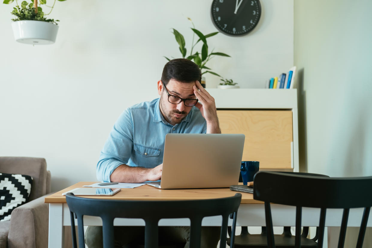 Worried displeased businessman reading bad news on his laptop computer while sitting at desk with a cup of coffee and digital tablet in the living room.