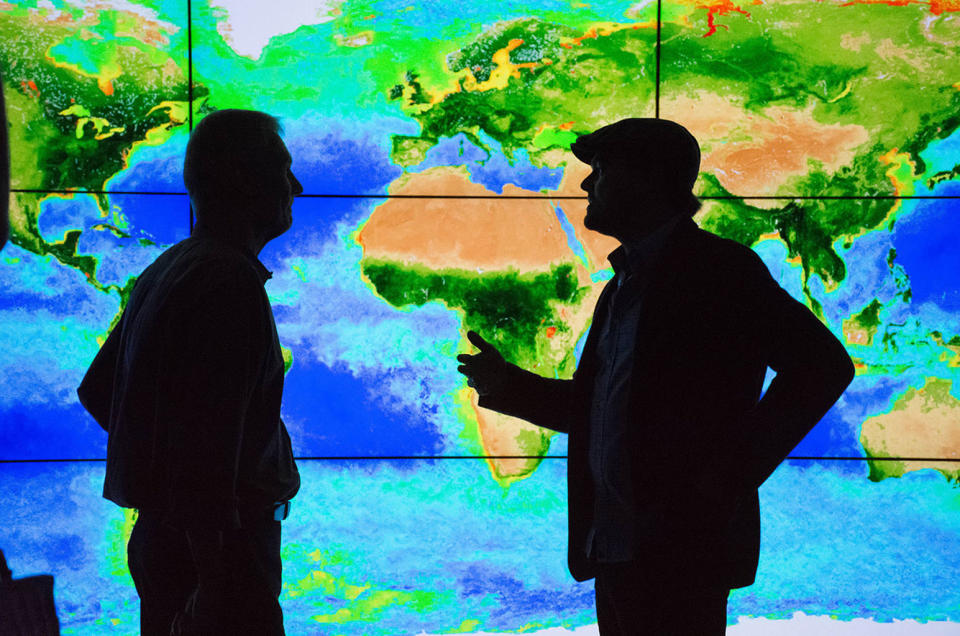 Actor Leonardo DiCaprio (right) speaks with Piers Sellers in front of a wall display showing biosphere data at NASA’s Goddard Space Flight Center in Maryland in April 2016. <cite>NASA</cite>