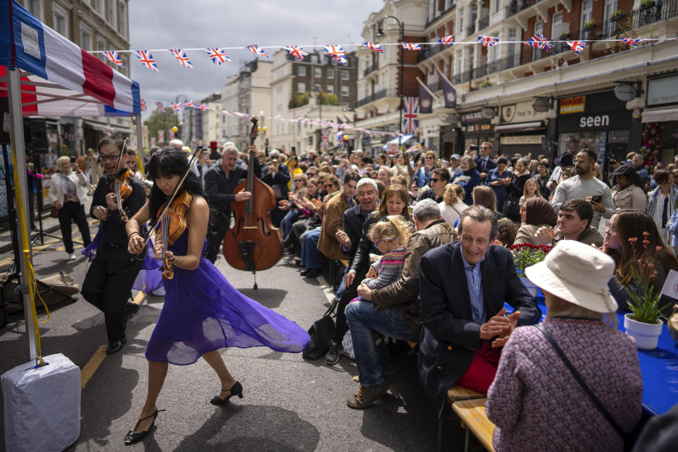 Musicians play their instruments as people sit at long tables to eat their lunch as part of the Big Lunch celebration in London, Sunday, May 7, 2023. The Big Lunch is part of the weekend of celebrations for the Coronation of King Charles III. (AP Photo/Emilio Morenatti)