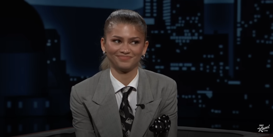 Woman in a grey suit with a tie, smiling, seated on a talk show set