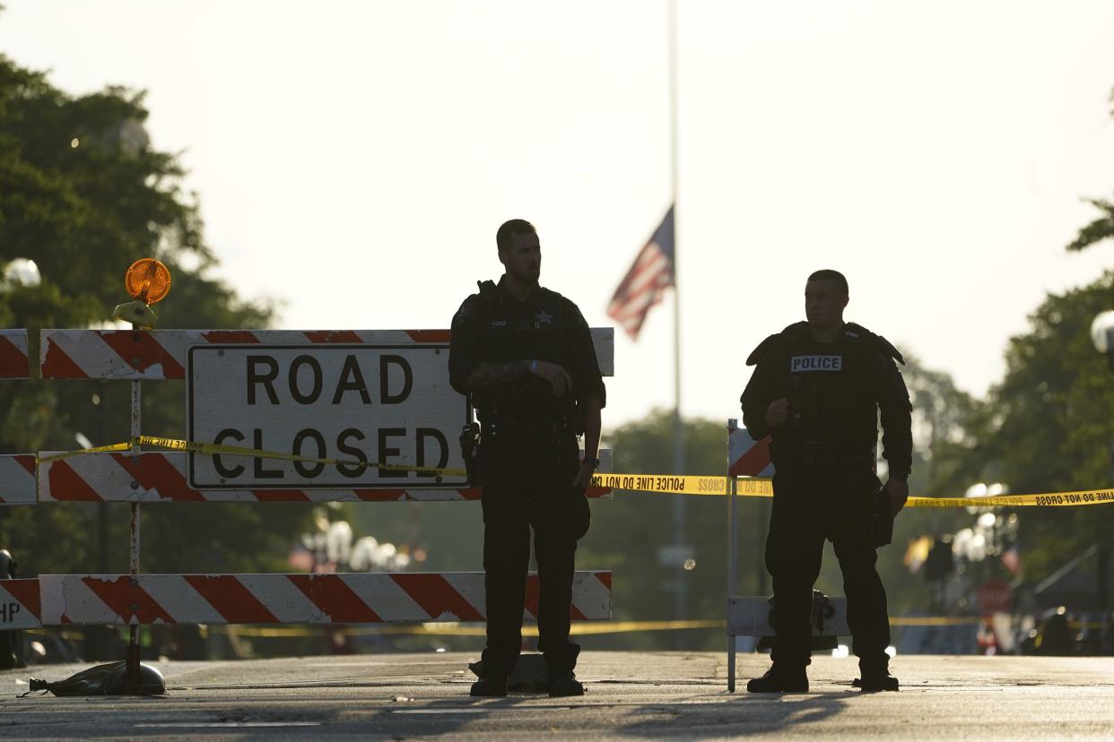 Two police officers stand their at post the day after a deadly mass shooting on the Westside of Highland Park, Ill. on Tuesday, July 5, 2022, as the American flag flies at half-staff on the Eastside. A shooter fired on an Independence Day parade from a rooftop, spraying the crowd with gunshots initially mistaken for fireworks before hundreds of panicked revelers of all ages fled in terror.
