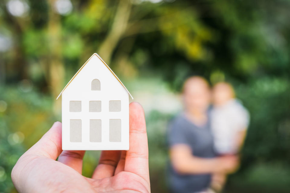 A hand holding a small house model in the foreground, with an out-of-focus person holding a child in the background