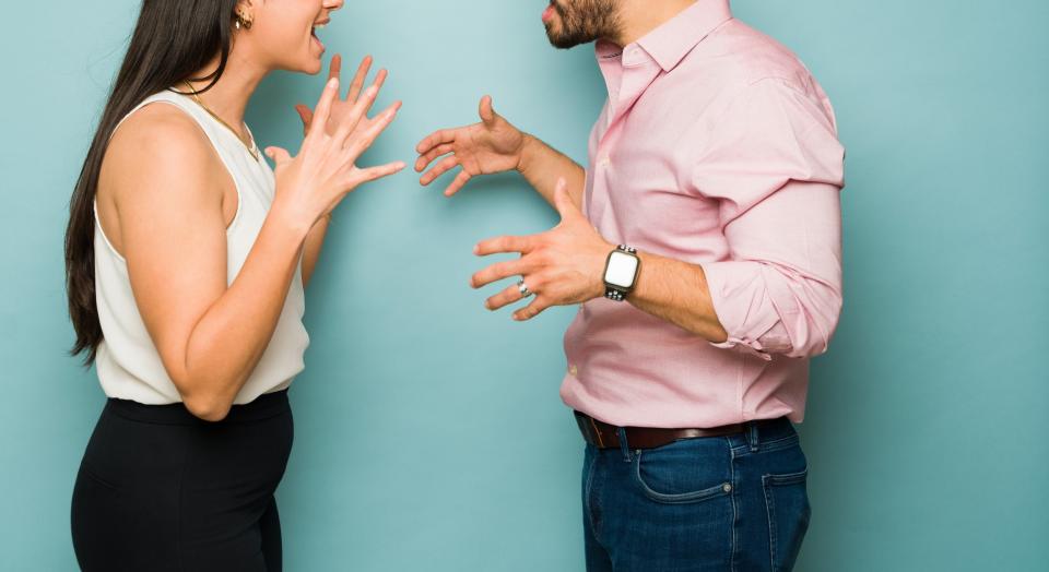 Man and woman face each other, both gesturing and appearing to argue. The man wears a pink shirt and jeans, the woman a white top and black pants