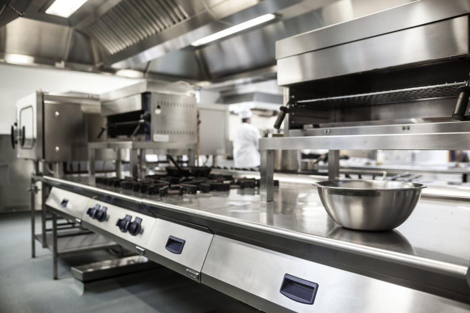 A professional, industrial kitchen with stainless steel appliances, cooking equipment, and a mixing bowl on the counter. A chef is working in the background