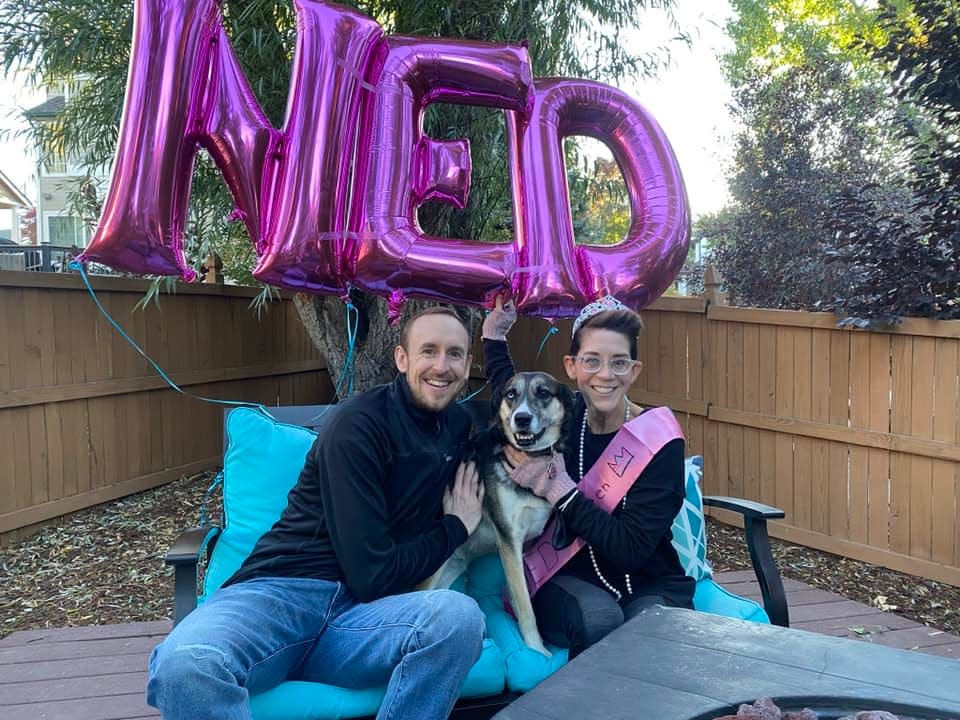 Katie wearing a tiara and holding balloons that say "NED" alongside Nick and their dog, Alice.