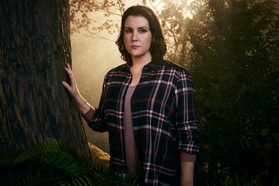 yellowjackets star melanie lynskey, in character as shauna, stands in a forest with her right hand resting on a tree trunk