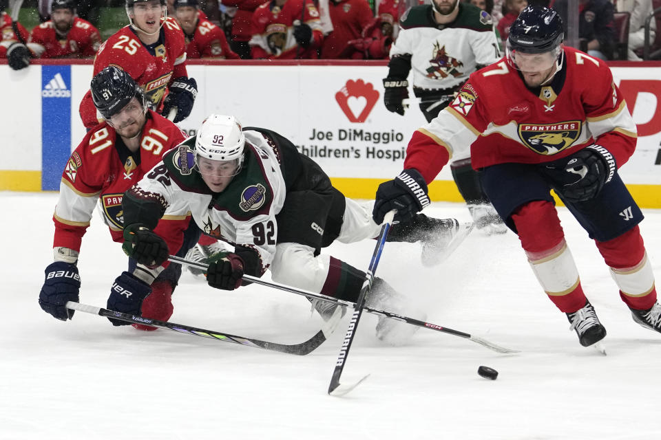 Arizona Coyotes center Logan Cooley (92) falls to the ice as he goes for the puck against Florida Panthers defensemen Oliver Ekman-Larsson (91) and Dmitry Kulikov (7) during the first period of an NHL hockey game, Wednesday, Jan. 24, 2024, in Sunrise, Fla. Ekman-Larsson was penalized for tripping on the play. (AP Photo/Lynne Sladky)