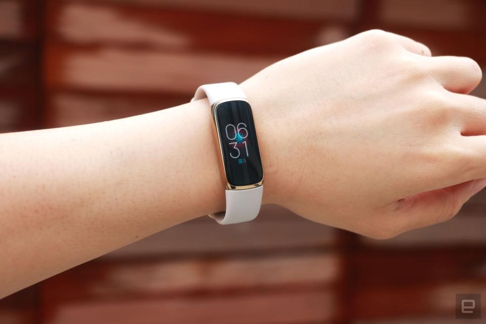 <p>Front view of the Fitbit Luxe with a light pink silicone band on a wrist against a dark brown paneled background. The screen shows the time is 6:31pm.</p>
