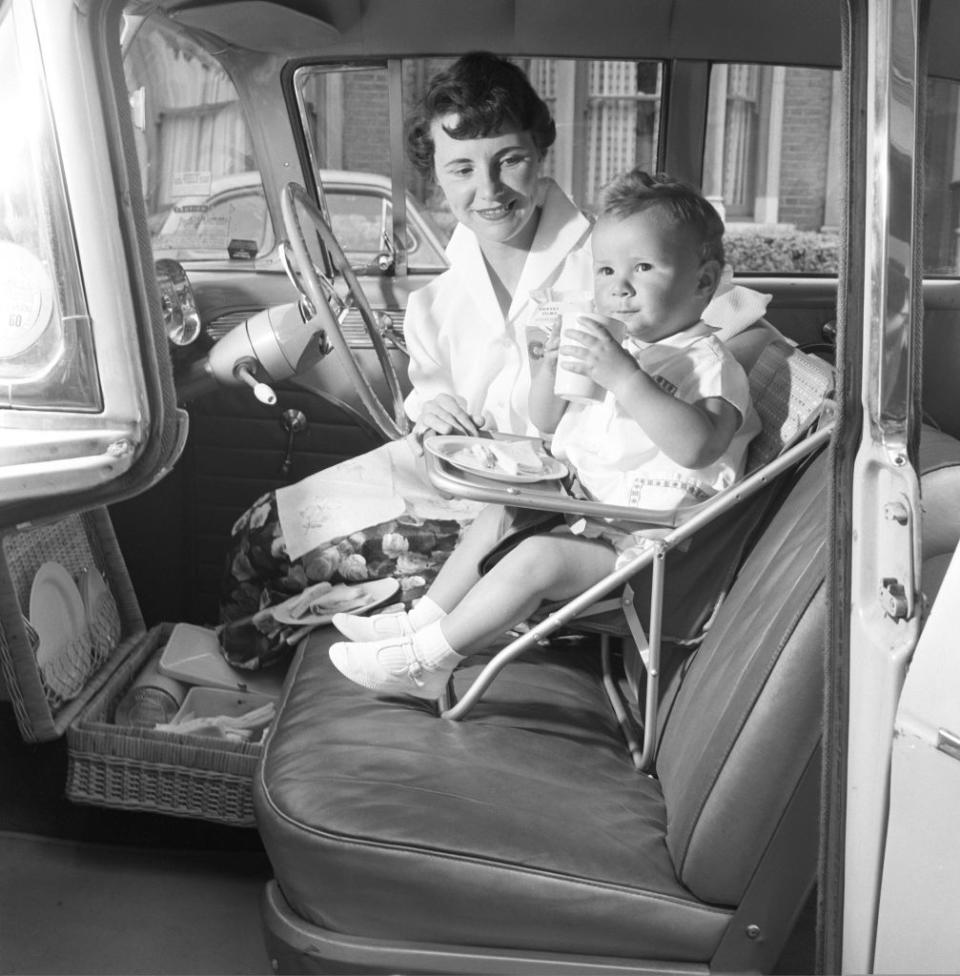 Woman feeding a child in a vintage car's front seat with tray