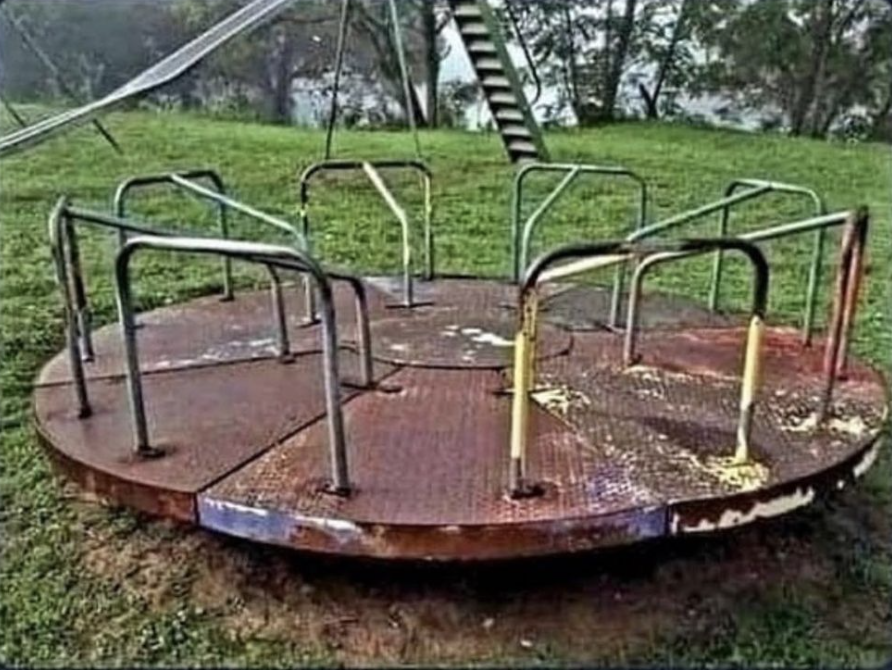 Old playground merry-go-round with chipped paint and rusty metal handles, placed in a grassy park area