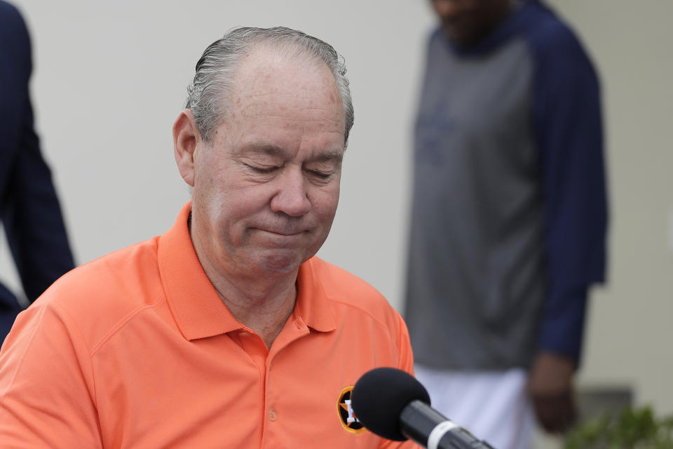 Jim Crane sits at a microphone in an orange shirts with a sad face.