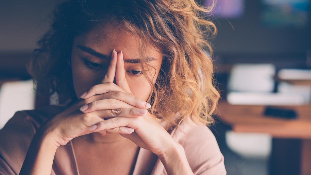 Closeup of sad young Asian woman at cafe leaning head on clasped hands and staring into vacancy.