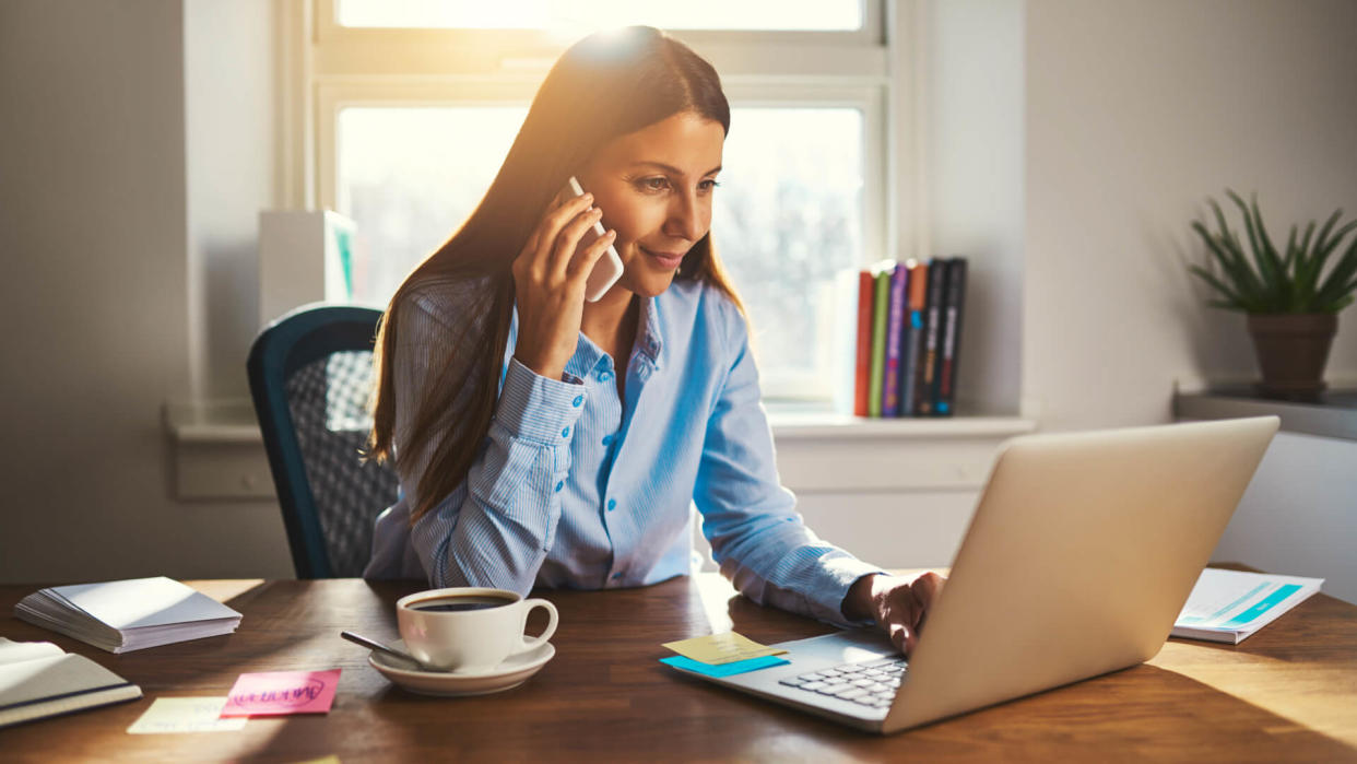 Woman working on laptop at office while talking on phone, backlit warm light.