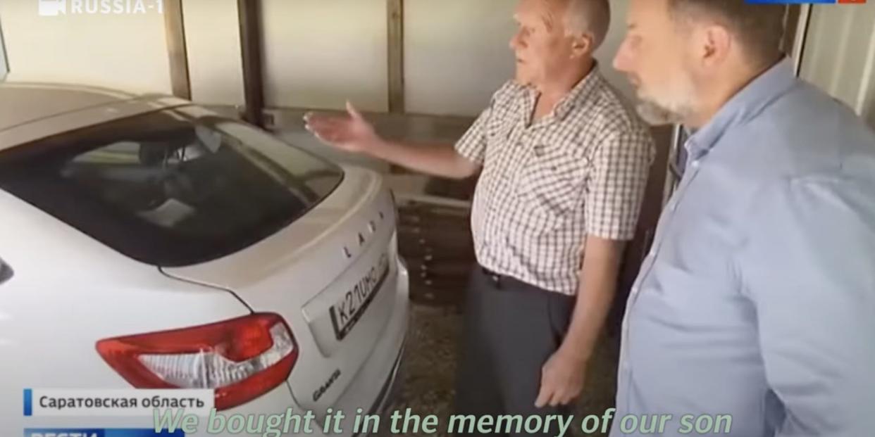 A bereaved Russian father shows a Russia-1 reporter a white Lada bought with "coffin money." Subtitles read: "We bought it in the memory of our son."