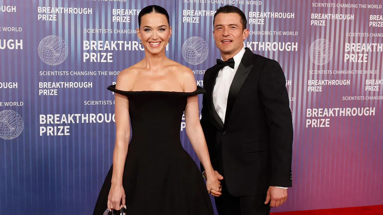 Katy Perry in a black off the shoulder dress holds hands with Orlando Bloom in a traditional tuxedo at the 2024 Breakthrough Prize Ceremony