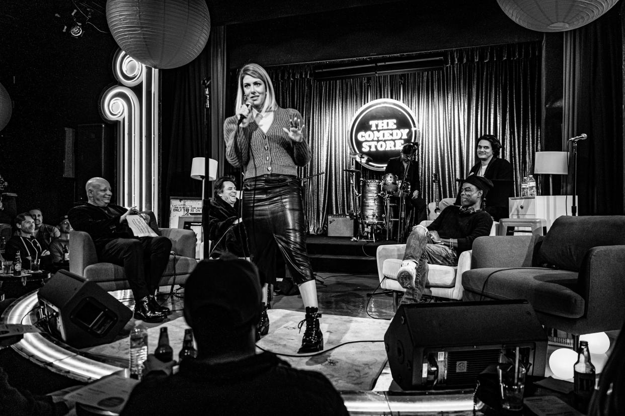 Kelly Rizzo-Saget in a leather midi skirt and button down cable knit sweater holds a microphone smiling onstage in front of Chris Rock, John Mayer and Jim Caery. (Mathieu Bitton/Netflix)