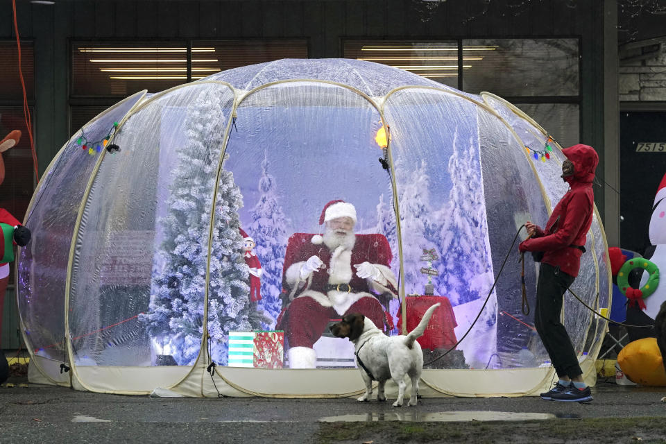 FILE - Santa Claus, portrayed by Dan Kemmis, laughs as he talks to Kristin Laidre as she walks her dog, Scooby, a Bassett Hound mix, as he sits inside a "snow globe" protective bubble due to the coronavirus pandemic in Seattle's Greenwood neighborhood on Dec. 8, 2020. (AP Photo/Ted S. Warren, File)