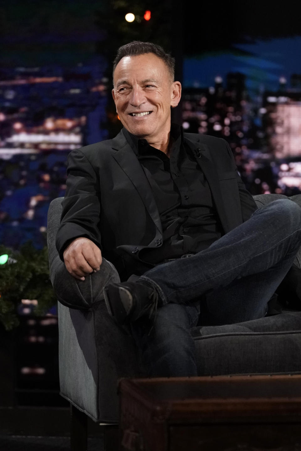 Célébrité masculine assise les jambes croisées dans un talk-show portant une chemise et une veste noires