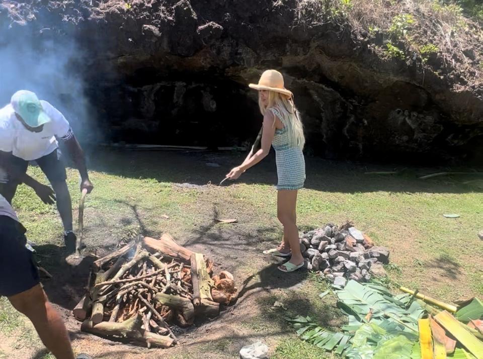 The writer wears a green and white dress and a sunhat and holds a piece of wood as two men in front of her start a campfire in a grassy area