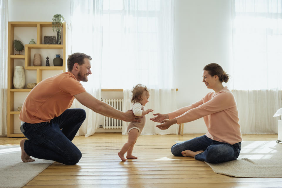 A man and woman encourage a baby to walk between them in a bright, cozy living room. Shelves with decorative items are in the background