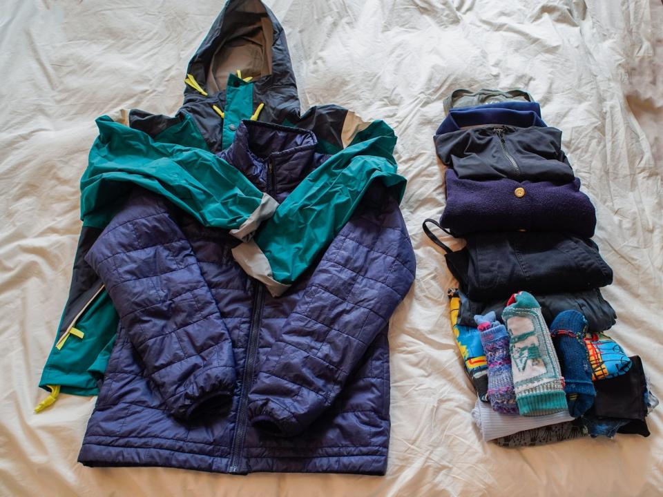 From left to right:  and a row of two rolled-up shirts and two fold-up long sleeve layers that are gray, black, navy blue folded teal pants, folded black pants, a folded blue and yellow t shirt, folded gray leggings, and a stack of four pairs of blue and black socks, a light blue tank top, and five pairs of dark-colored underwear, all on a white sheet