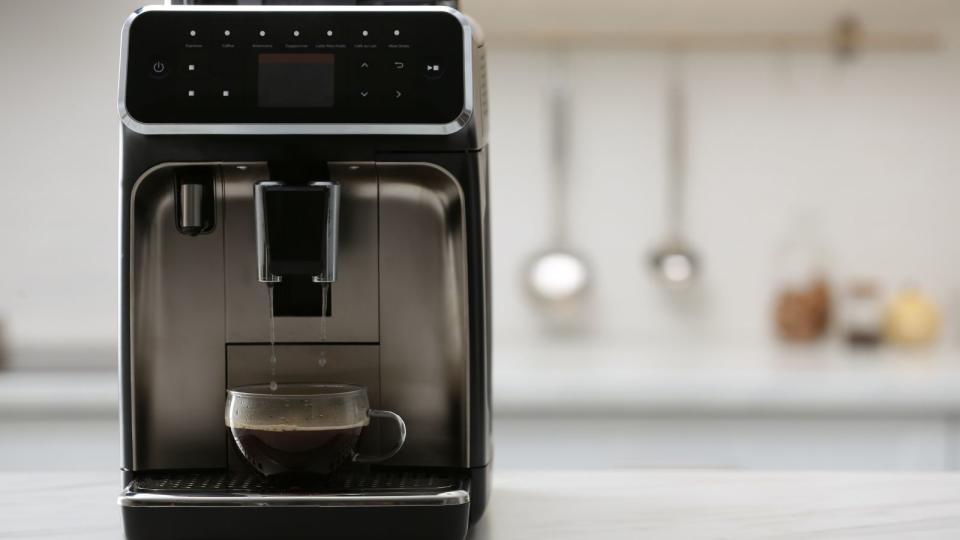 A black coffee maker on a countertop with a  blurred kitchen in the  background