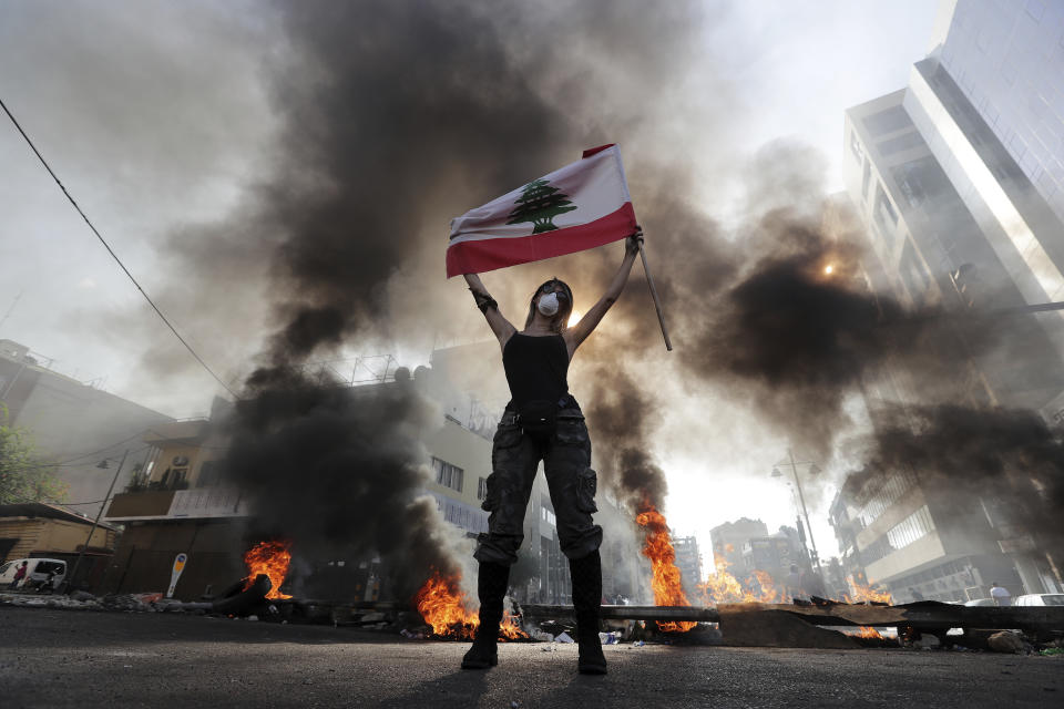 An anti-government protester holds a Lebanese flag while standing in front burning tires that block a road in the town of Jal el-Dib, north of Beirut, Lebanon, Wednesday, Nov. 13, 2019. Lebanese protesters blocked major highways with burning tires and roadblocks on Wednesday, saying they will remain in the streets despite the president's appeal for them to go home. (AP Photo/Hassan Ammar)