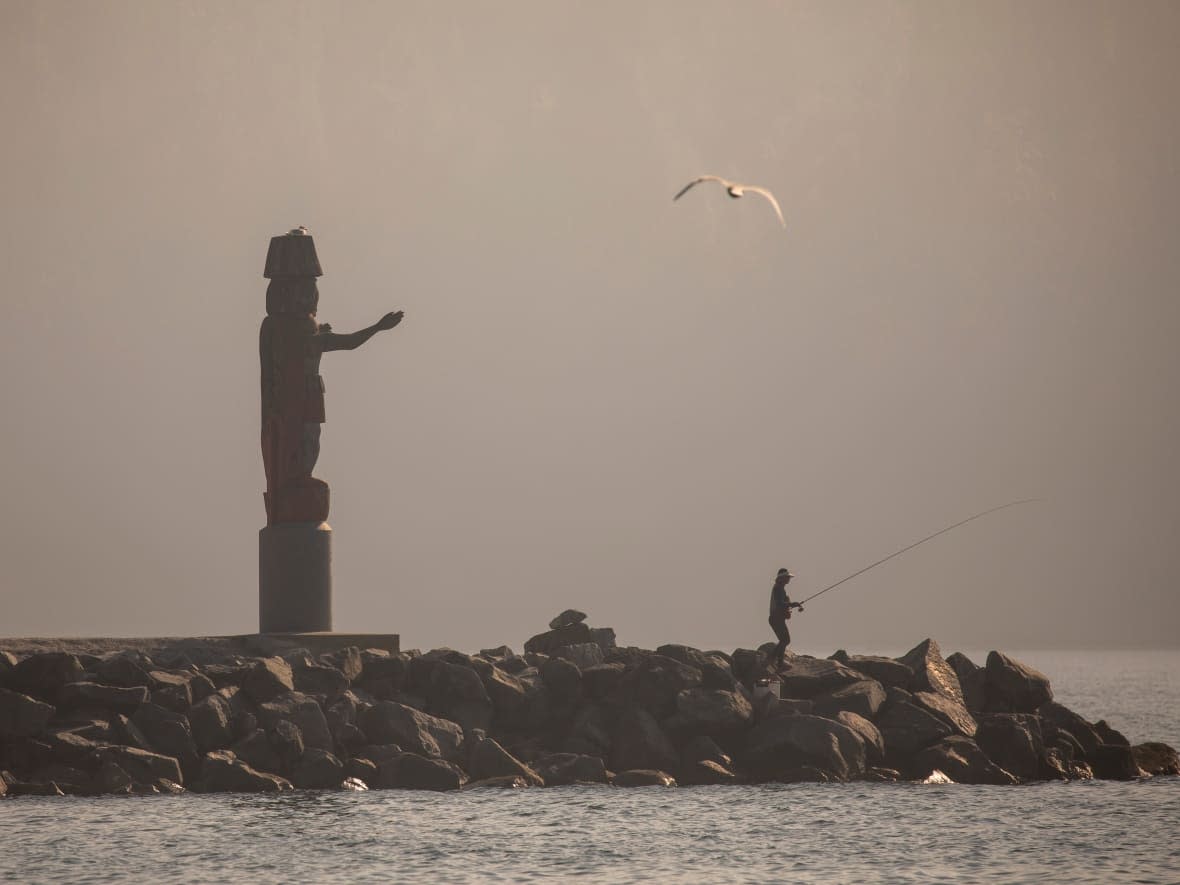 Smoky skies on Wednesday over Metro Vancouver blur the Squamish Nation welcome figure and a lone person fishing at Ambleside Beach in West Vancouver, B.C. (Ben Nelms/CBC News - image credit)