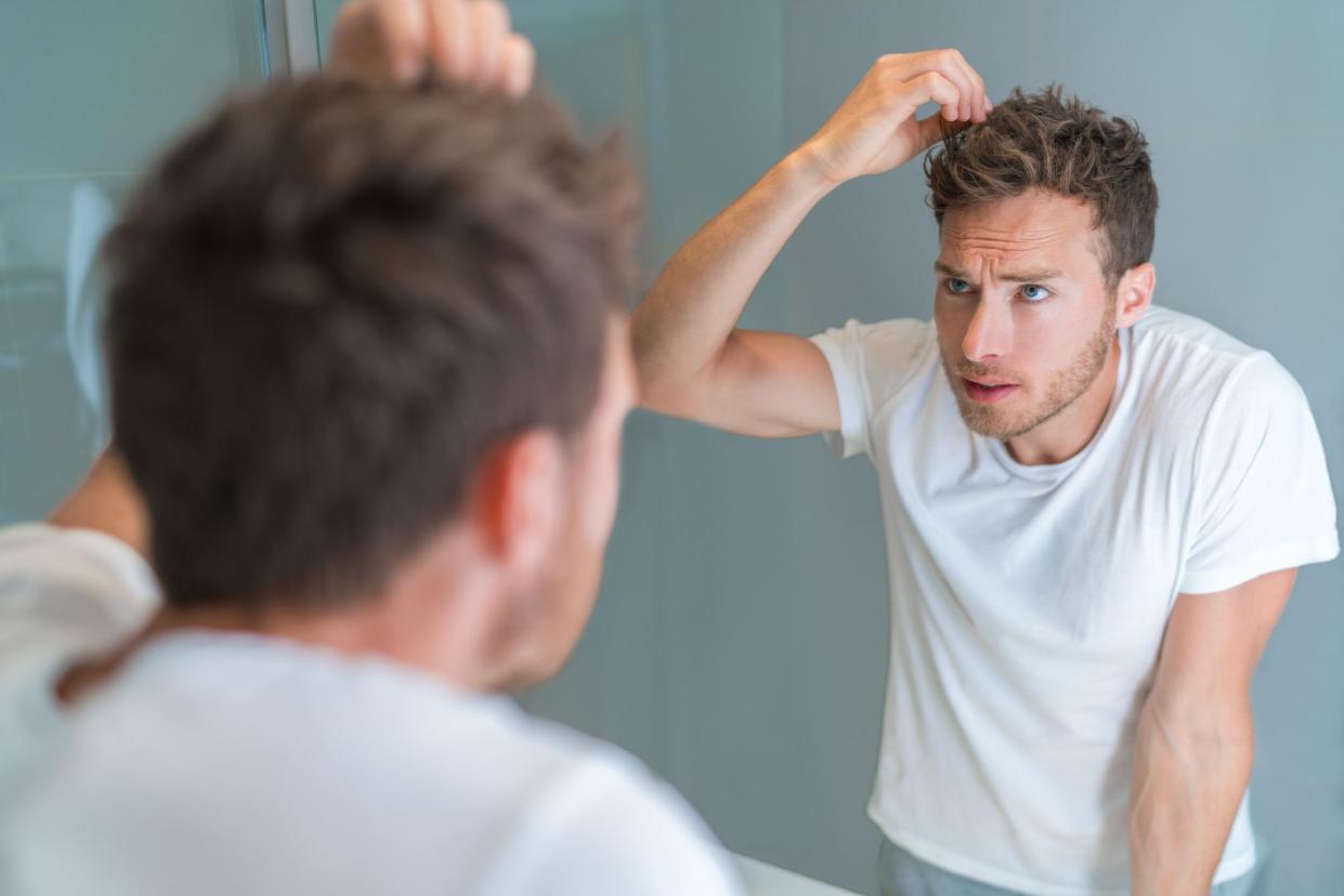 Hair loss man looking in bathroom mirror styling hairstyle with gel or checking for hair loss or grey hairs. Unhappy male health problem
