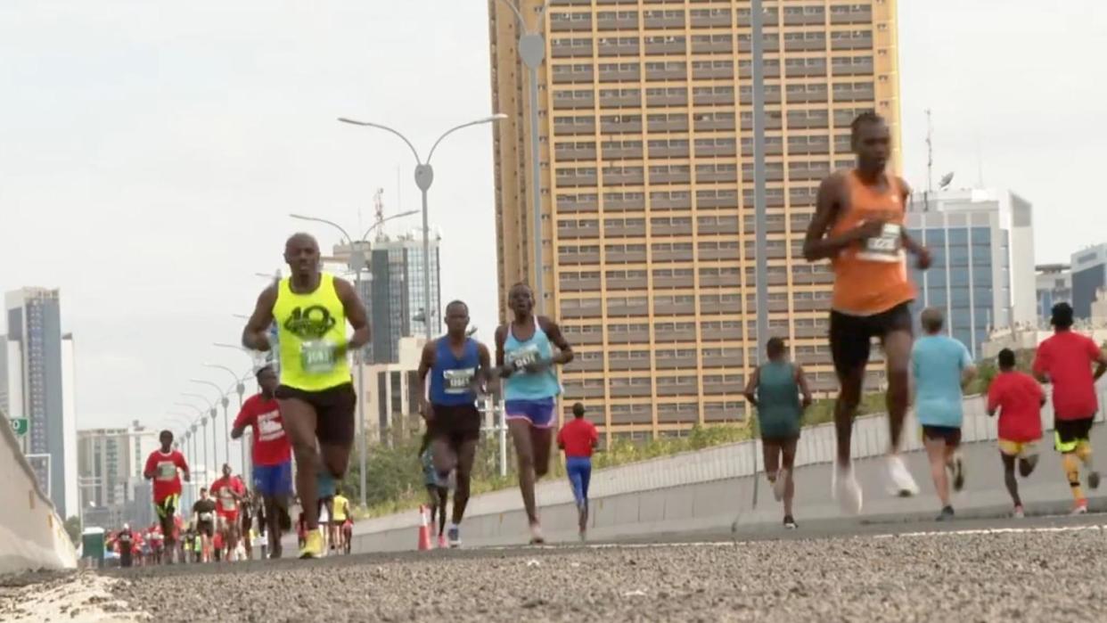 Runners follow a course along a road in Kenya with a backdrop of high-rise buildings