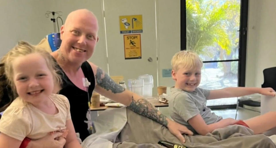 Harry Slabbers can be seen in a hospital bed smiling beside his son and daughter beside him, putting on a brave face despite the extra challenges Perth's rental crisis is putting on his cancer journey.