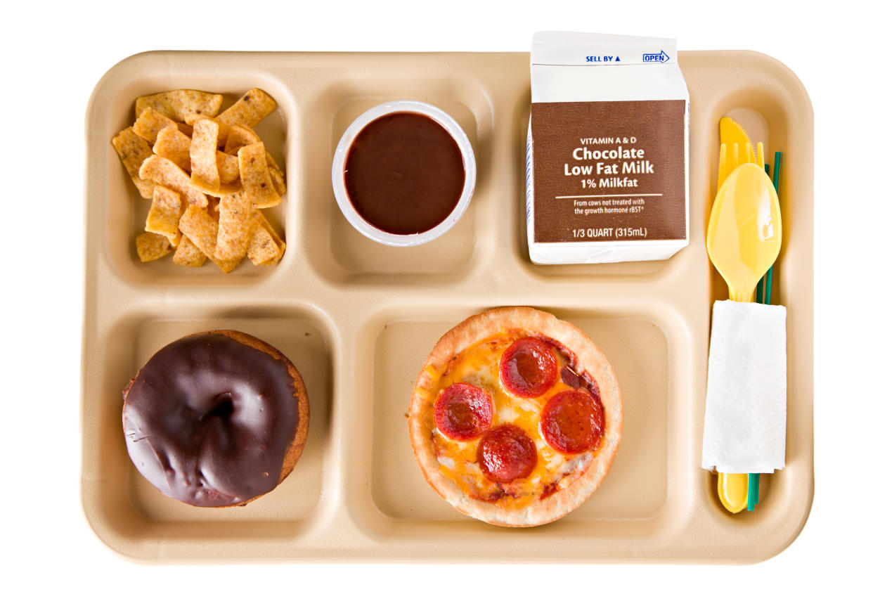 A school lunch tray contains a doughnut, a mini pepperoni pizza, corn chips, pudding and low-fat chocolate milk.