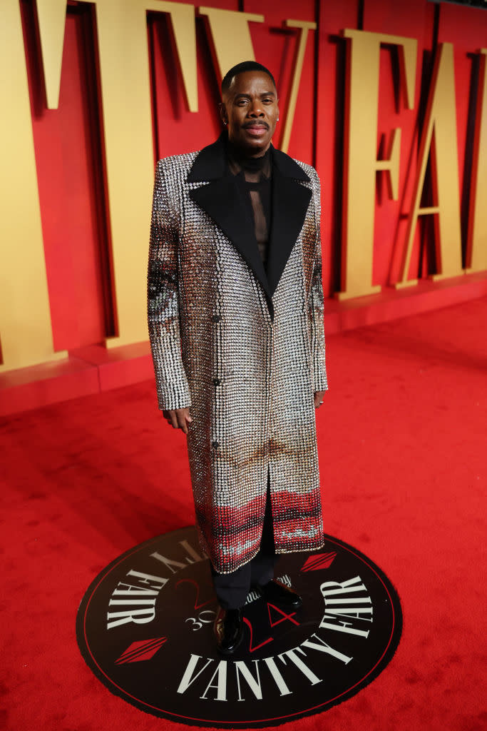 A person in a sequined coat and trousers standing on a Vanity Fair event carpet