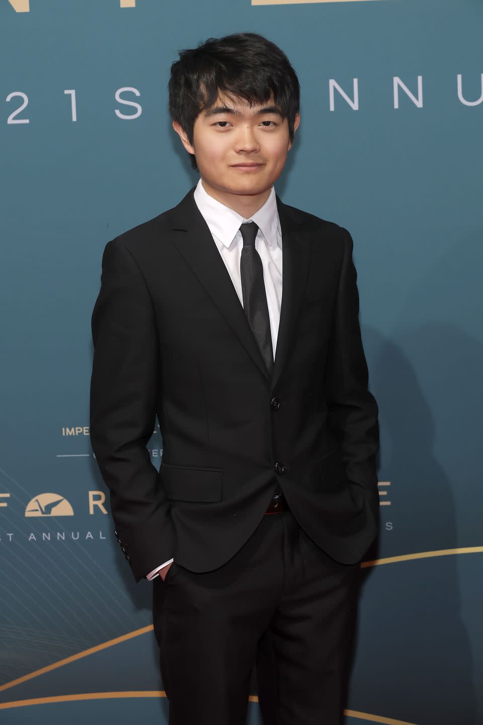 ben wang poses on the red carpet wearing a black suit with a white shirt and black tie, smiles at the cameras with his hands in his pockets