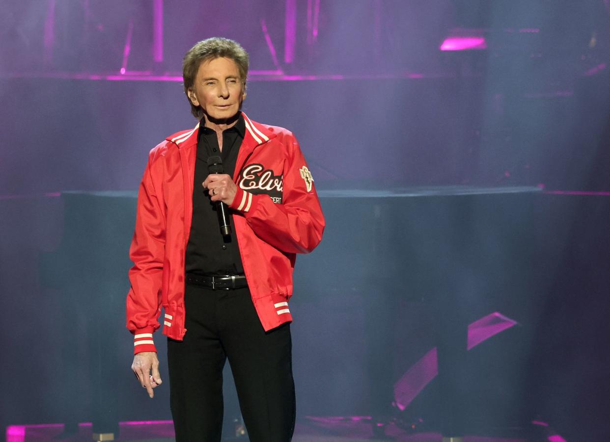 barry manilow stands on a stage in a black shirt and pants with a red jacket