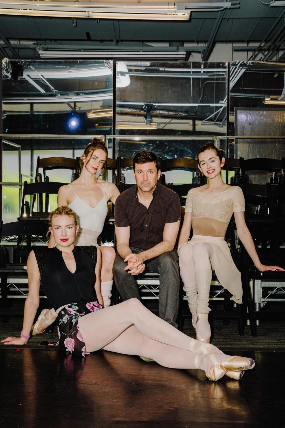 A man in street clothes sits with three ballerinas in rehearsal clothes in a studio