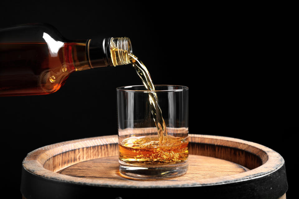A bottle is pouring whiskey into a glass on a wooden barrel with a black background