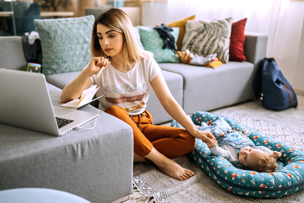 A woman holds hands with her infant while looking for something on her laptop. 