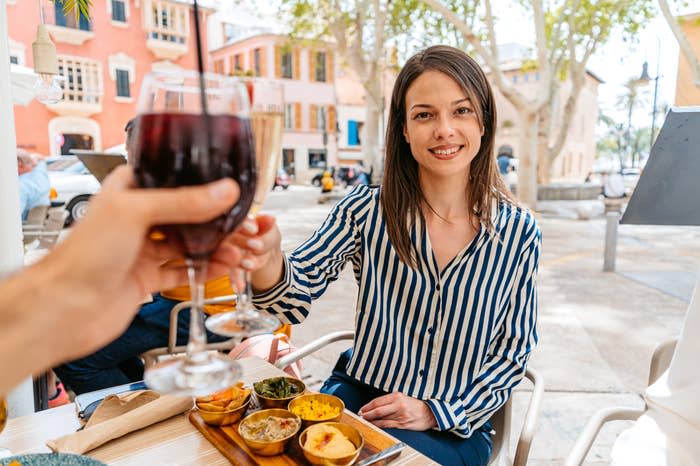 A woman in a striped blouse enjoys a meal and raises a glass of wine in a toast at an outdoor restaurant. Another person's hand holding a glass is in the foreground