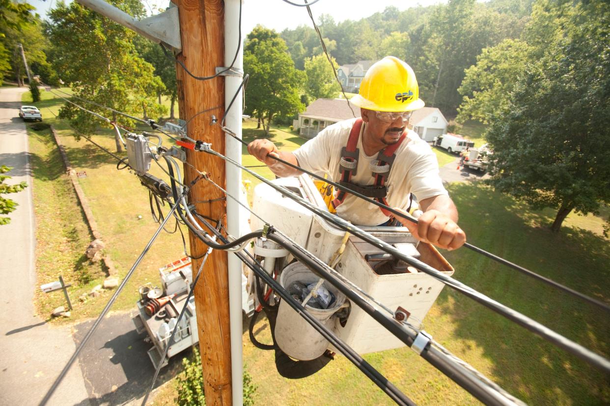 utility worker wearing safety goggles and yellow hard hat standing in a cherry picker holds onto power line cable next to utility pole above a green lawn in front of house among green trees