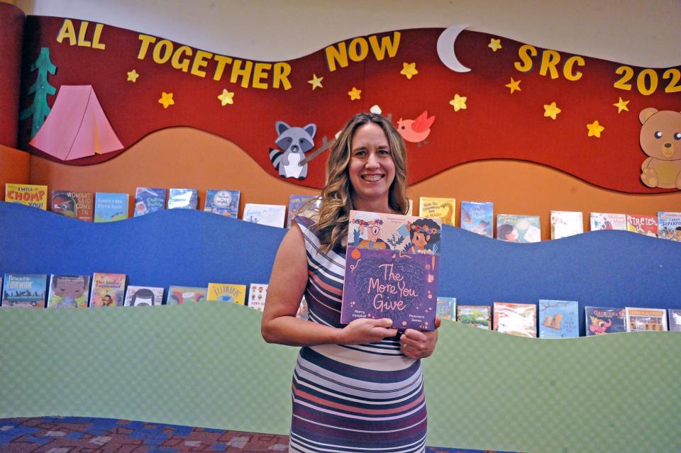 Author Marcie Campbell stands with her latest published children's book, The More You Give, in front a rack of books in the children's department at the Wayne County Library main branch in Wooster.