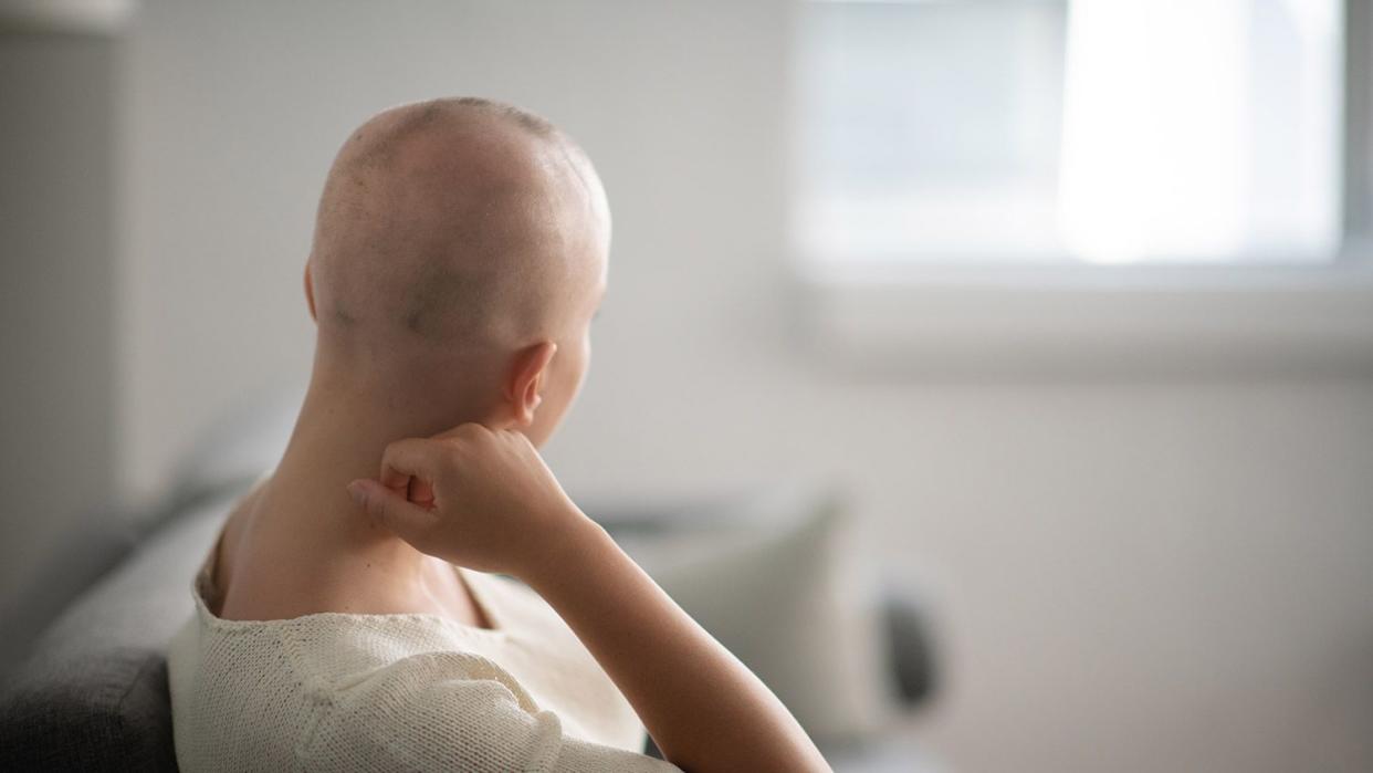 A woman is indoors in her living room. Her head is shaved due to chemotherapy. She is sitting and looking thoughtful.