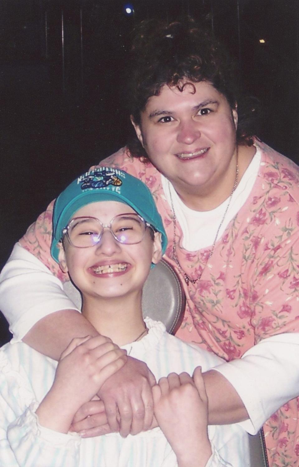 dee dee blanchard holding her arms around gypsy rose blanchard, who is smiling widely, sitting in a chair, and wearing a hat on her head