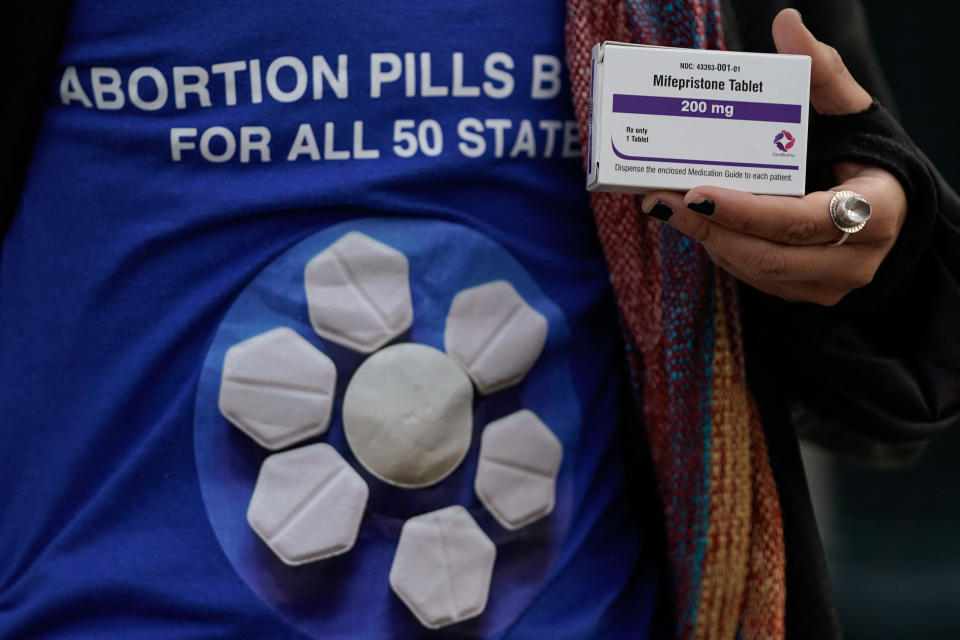 Person wearing a shirt with "Abortion Pills For All 50 States" holding a Mifepristone tablet pack