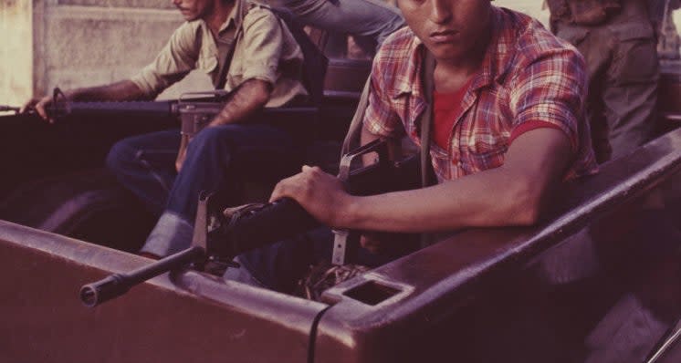 Three individuals in a pickup truck with one holding a rifle; civilians and soldiers visible in the background